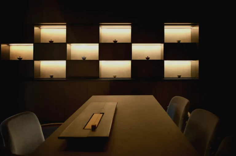 Tea Room by Ki-setsu, a destination of luxury chinese tea appreciation in Singapore. A dimly lit room with a wooden table and chairs in the foreground. Illuminated shelves on the wall display small bowls, creating a serene, minimalist ambiance.