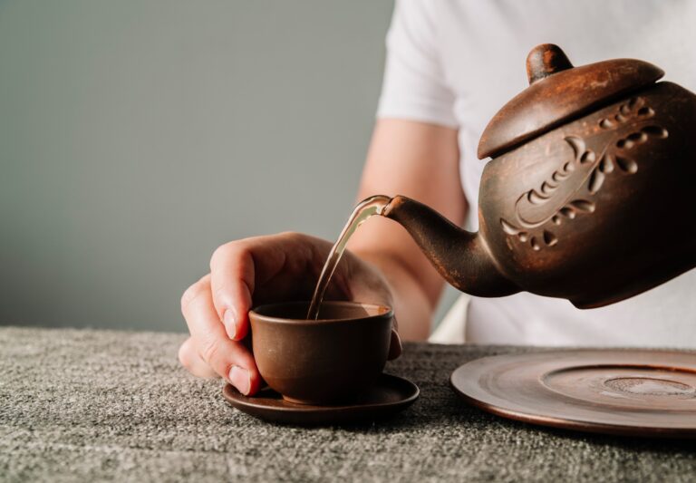 A hand gently pours hot tea from a traditional Chinese teapot into a small porcelain cup, capturing the calm, flowing motion of a classic tea ceremony.