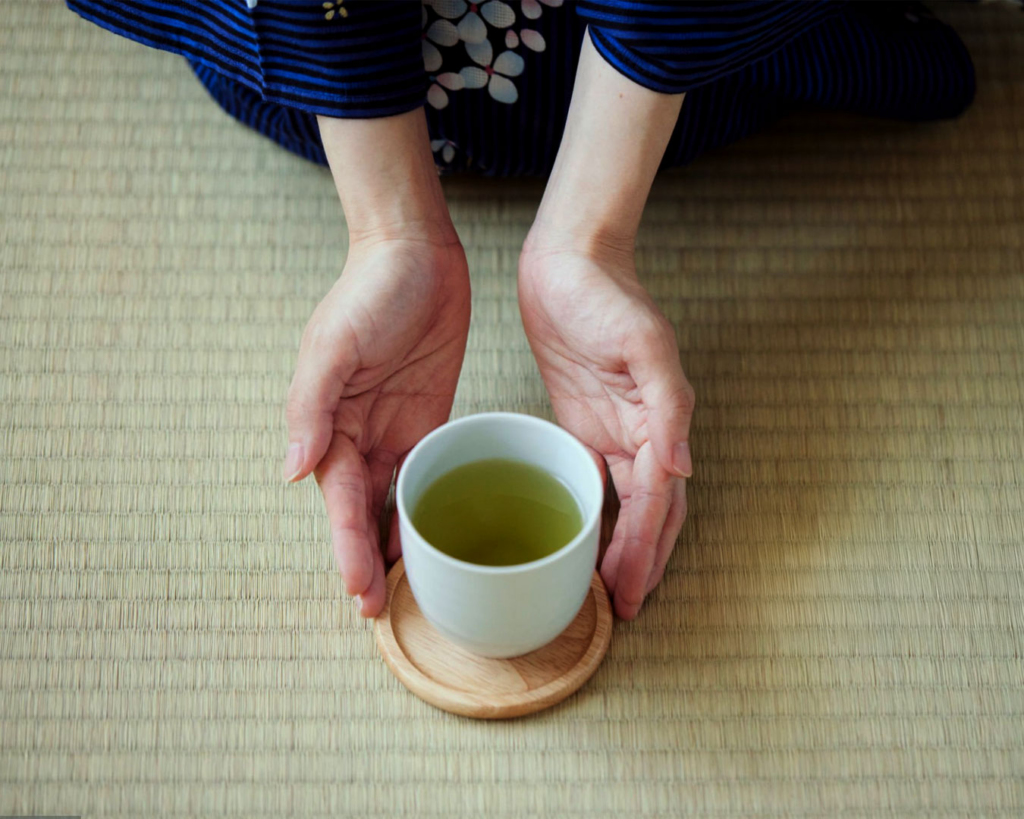 Hands offering a freshly brewed cup of green tea, symbolising hospitality, warmth, and the act of sharing a peaceful tea moment.