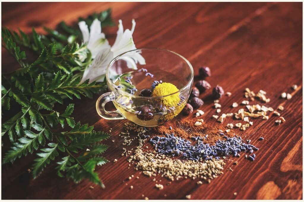 Glass teacup with herbs, lavender, and spices on a wooden table. Nearby are white lilies and green leaves, creating an earthy, calming atmosphere.