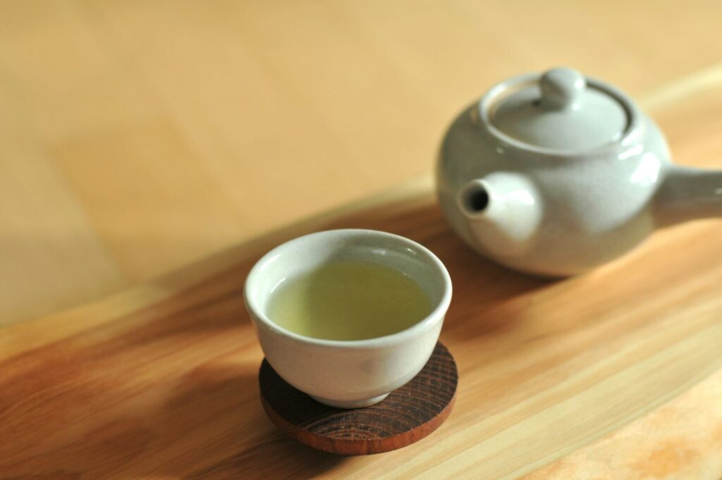 A serene setting with a pale ceramic teacup filled with green tea on a dark wooden coaster next to a compact teapot on a light wooden surface.