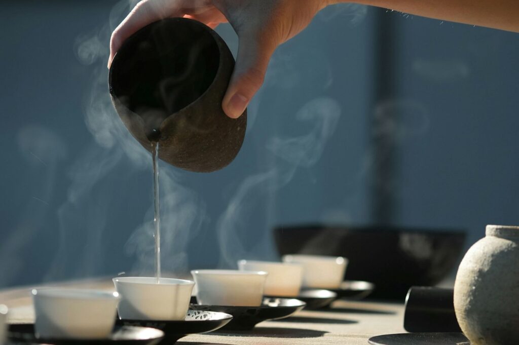 A hand pours steaming tea from a dark ceramic pot into a row of small white cups, creating a calm and serene atmosphere on a sunny day.