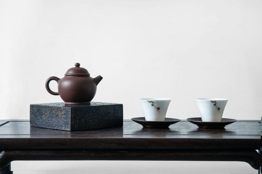 A brown teapot on a textured stand, flanked by two white cups with floral designs on saucers, sits on a dark wooden table against a plain backdrop.