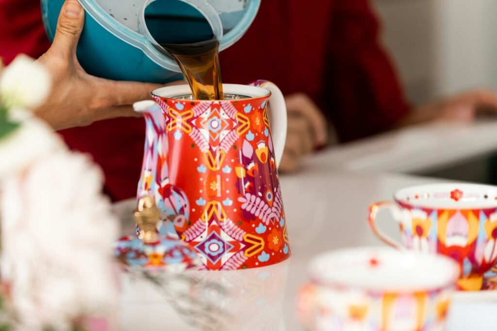 Person in red pouring coffee from a turquoise pot into a vibrant red and orange patterned teapot on a white table, with matching cups nearby.