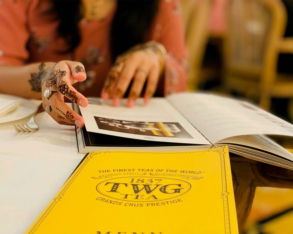 A woman seated at a table with a cup of TWG tea, calmly reading the menu in a tea lounge setting, creating a relaxed and refined tea-time atmosphere.