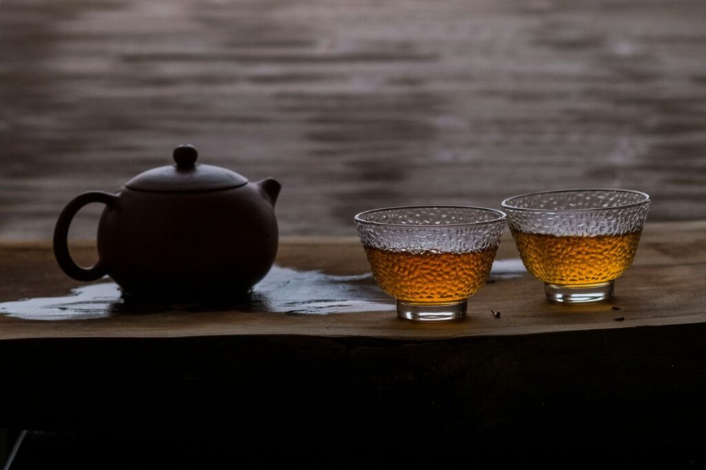 A dark brown teapot and two textured glass cups filled with amber tea sit on a wooden surface, conveying a serene and warm atmosphere.