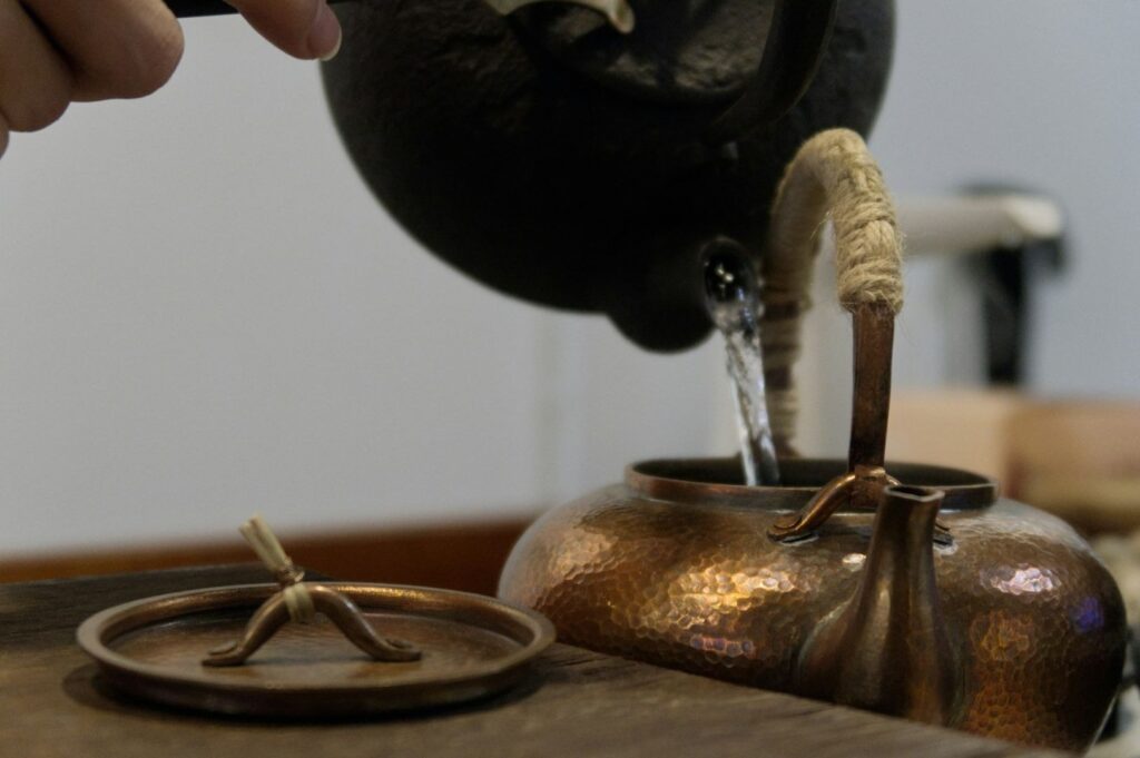 A hand pours water from a black kettle into a textured copper teapot with a rope-wrapped handle. The teapot lid lies beside it on a wooden surface.