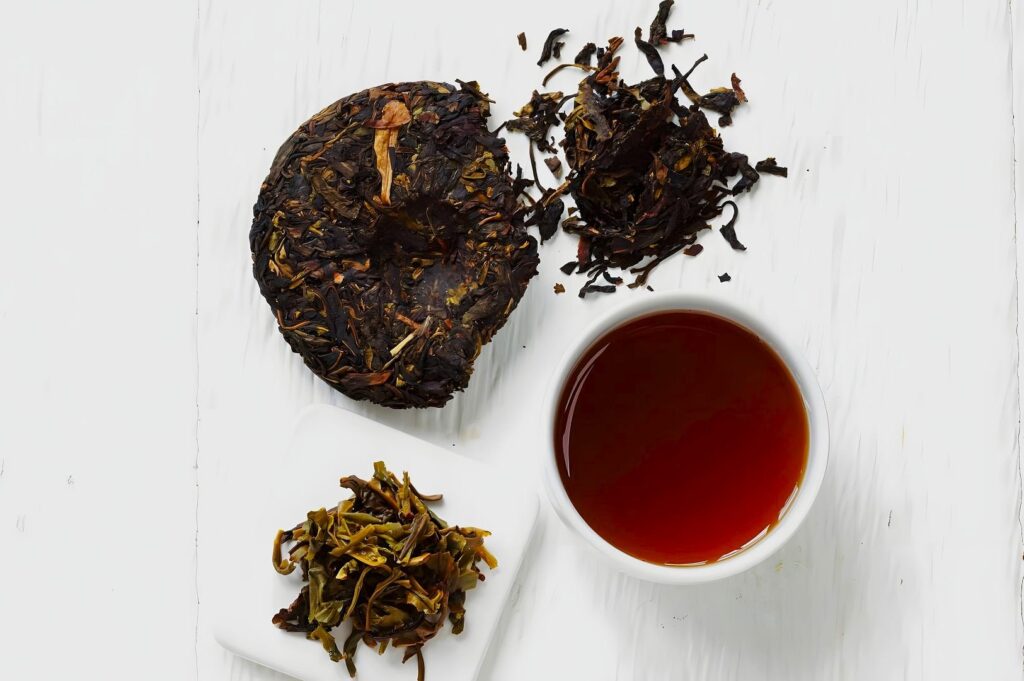 A round block of compressed tea, loose tea leaves, and a cup of brewed tea on a white background, conveying a fresh and earthy aroma.