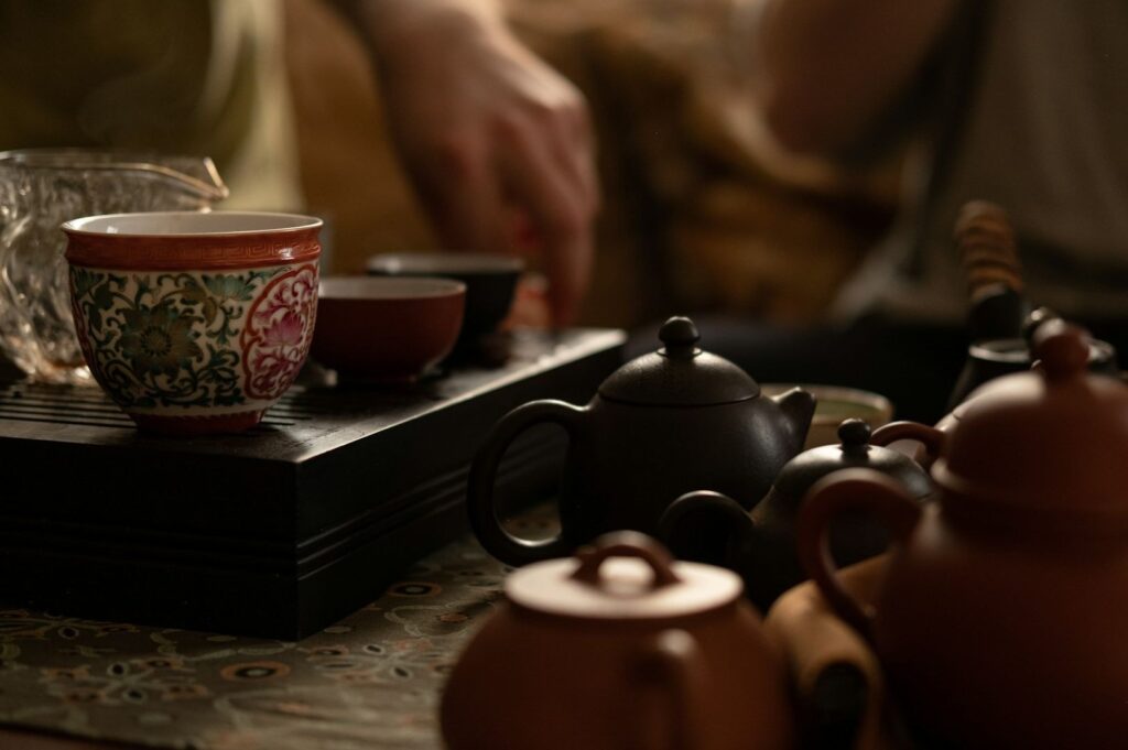Close-up of a traditional tea set with ornate cups and small teapots on a patterned tray. A hand reaches over, suggesting a peaceful tea ritual.