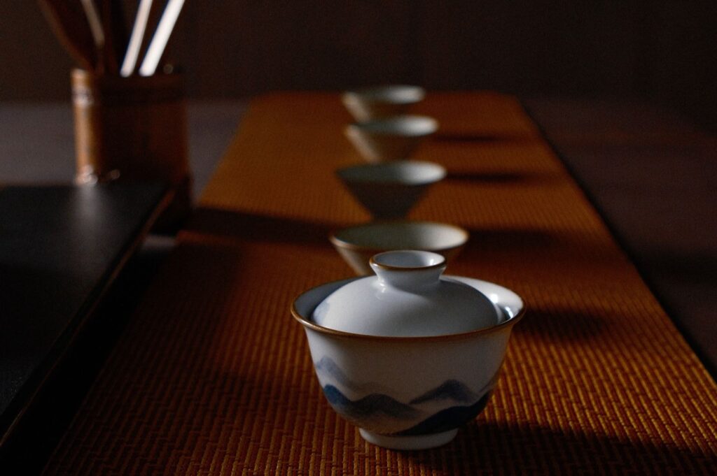 A row of elegant, white teacups with blue patterns is arranged on an orange mat, evoking a serene, minimalist ambiance. Soft lighting enhances the scene.