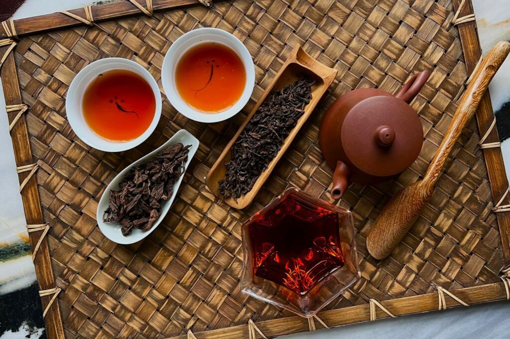 Tea set on woven mat with two cups of amber tea, dried leaves in wooden trays, a brown teapot, wooden stirrer, and a glass star-shaped cup. Calm and inviting.