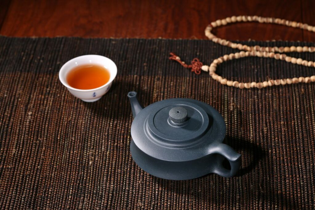 A black teapot and a cup of tea sit on a woven mat, next to a string of wooden beads. The warm lighting creates a calm, serene atmosphere.