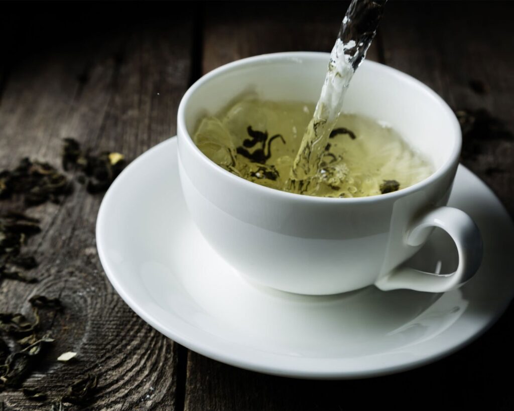 Fresh hot water flows into a clear teacup containing tea leaves, highlighting their movement as they unfurl. The scene captures the quiet moment of tea infusion.