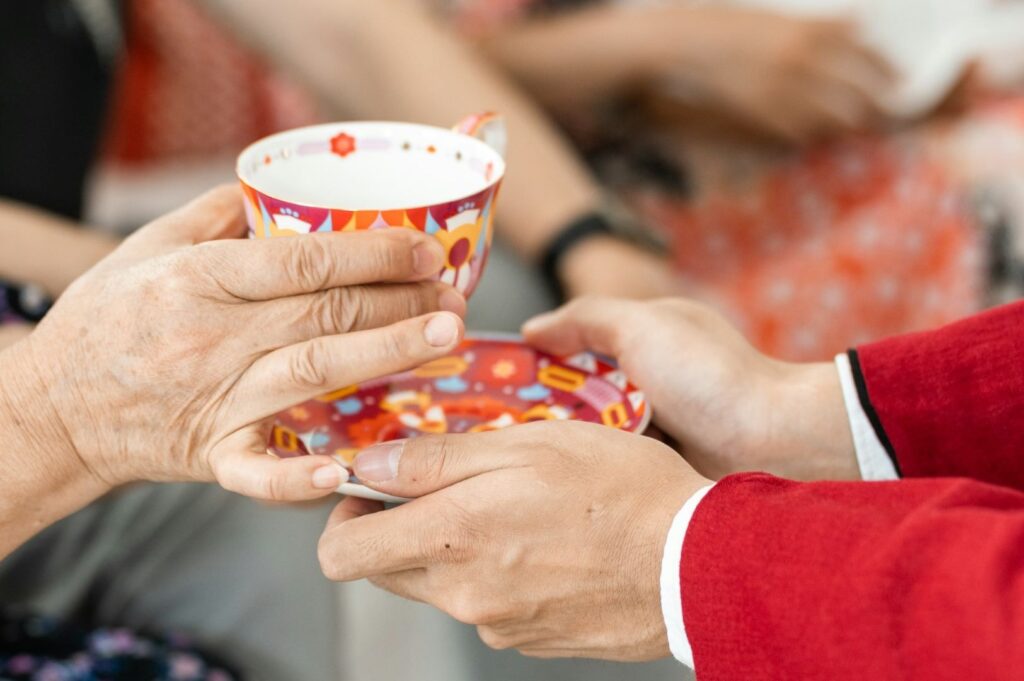 Two people exchange a colorful teacup and saucer. One person wears a red sleeve, while the other's hands appear older, suggesting a warm, caring interaction.