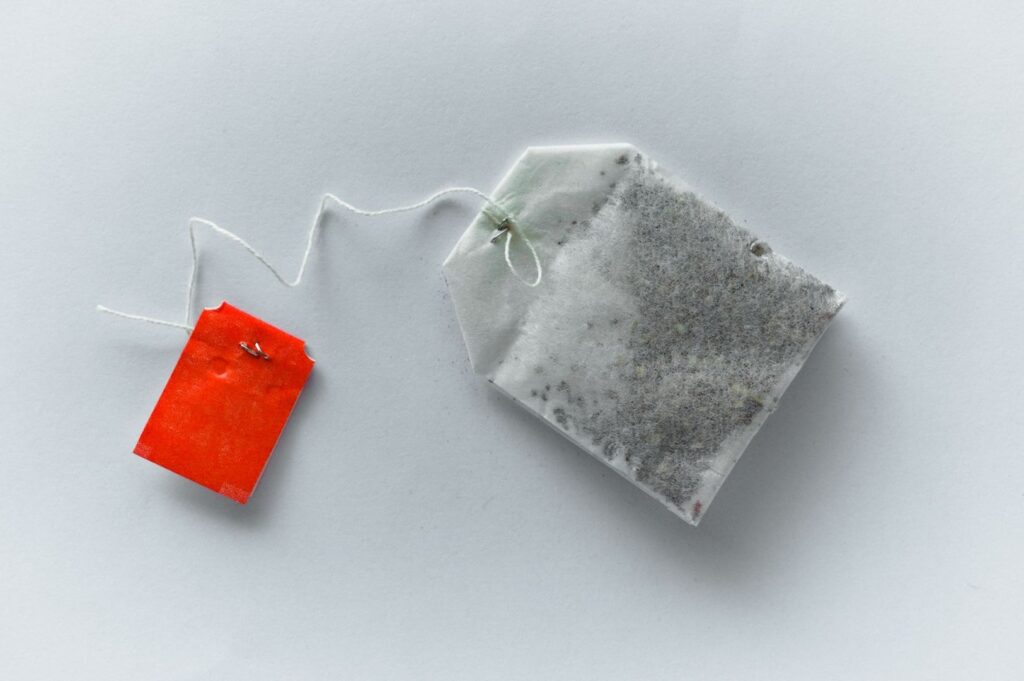 A white tea bag with visible tea leaves inside is attached to a string with a small red tag. The image has a minimalist, clean background.
