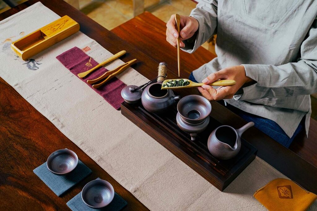 A person prepares tea with a traditional set on a wooden table. The scene includes a teapot, cups, and utensils, evoking a calm, ritualistic atmosphere.