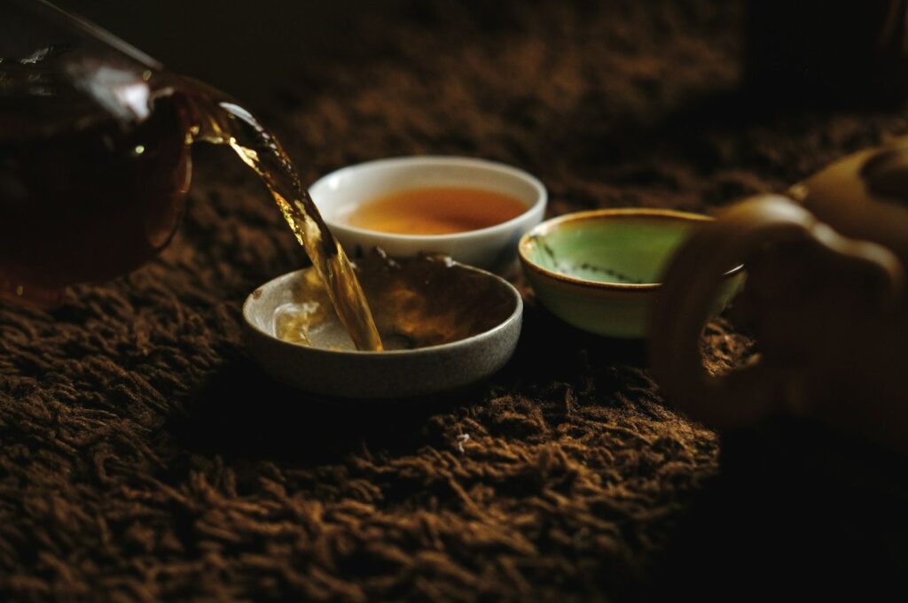 Tea is being poured into a small, rustic bowl on a textured brown surface, with two teacups nearby. The scene feels warm and calming.