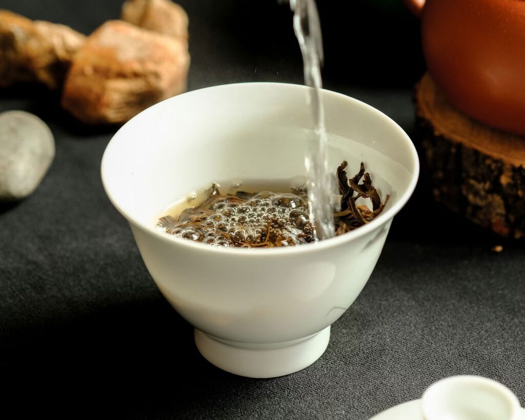 Hot water is poured into a teacup filled with loose tea leaves, allowing the leaves to slowly open and release their colour. Steam rises gently, showing the start of the brewing process.