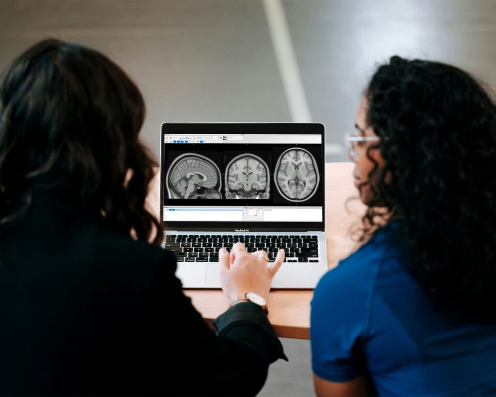 Two medical professionals studying a laptop screen displaying a human brain scan, suggesting scientific research or analysis related to health, cognition, or the effects of tea on the brain.