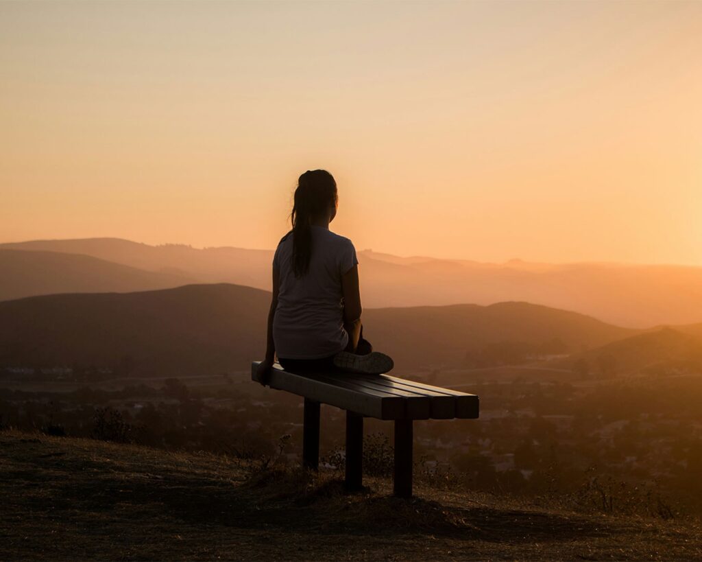 A woman seated on a tree branch, gazing at the sunset over the horizon, surrounded by warm orange light, expressing stillness, reflection, and mindful appreciation of nature.