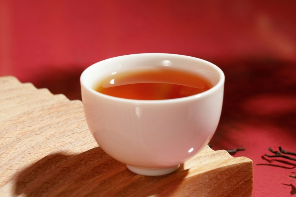 A small white cup filled with amber-colored tea sits on a light wooden surface against a soft red background, creating a warm and inviting atmosphere.
