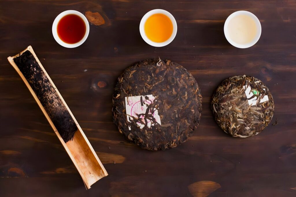 Three types of brewed tea in white cups are lined up above pressed tea cakes and a bamboo leaf, on a dark wooden surface. Earthy and rustic.