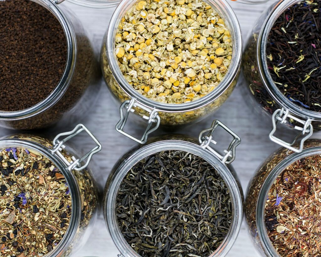 A visual display of six glass jars, each holding a different tea leaf variety, showcasing the natural progression of colour, texture, and processing styles across the tea spectrum.