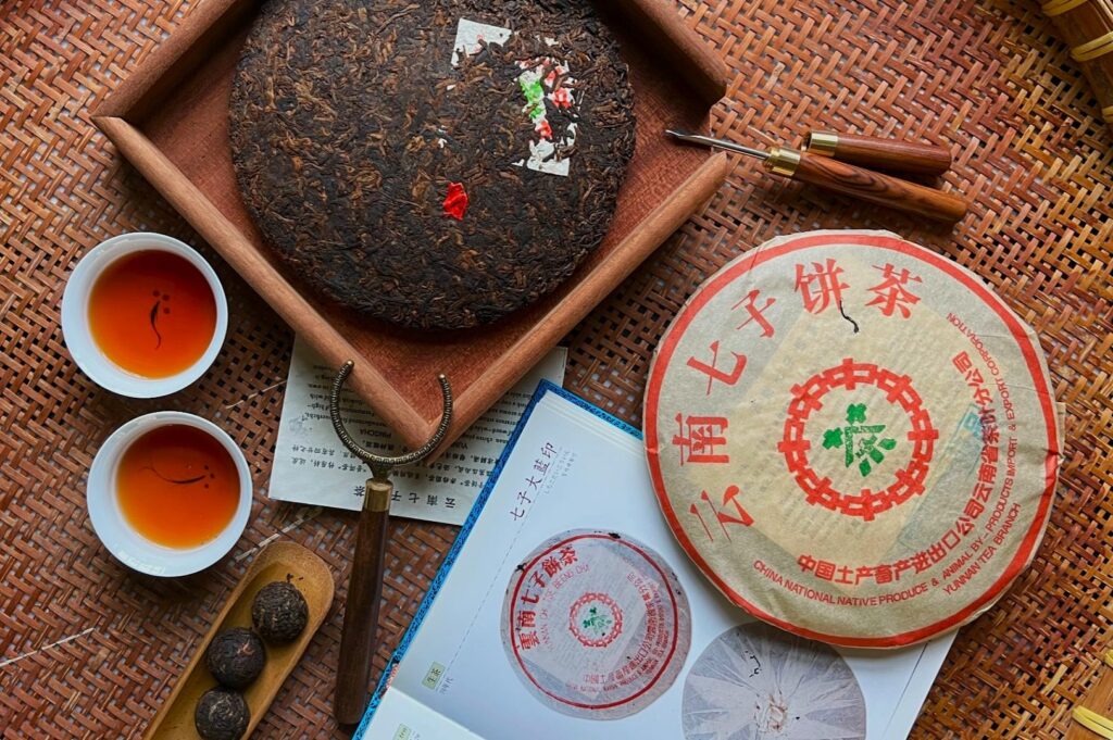 A round block of dark compressed tea sits on a wooden tray with brewing tools. Nearby, two cups hold amber tea. A wrapped tea package and a book lie beside them on a wicker surface. The scene conveys a sense of tradition and calm.