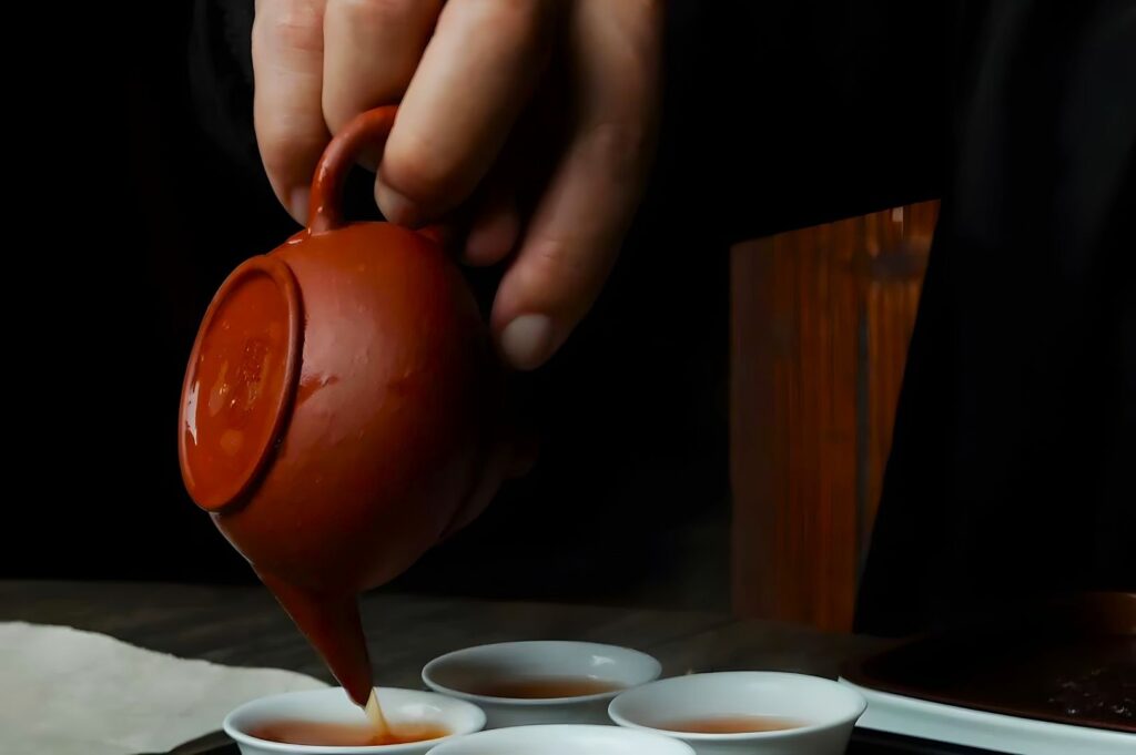 A hand pours tea from a small, clay teapot into white cups on a wooden table. The scene feels calming and traditional, emphasizing careful attention.