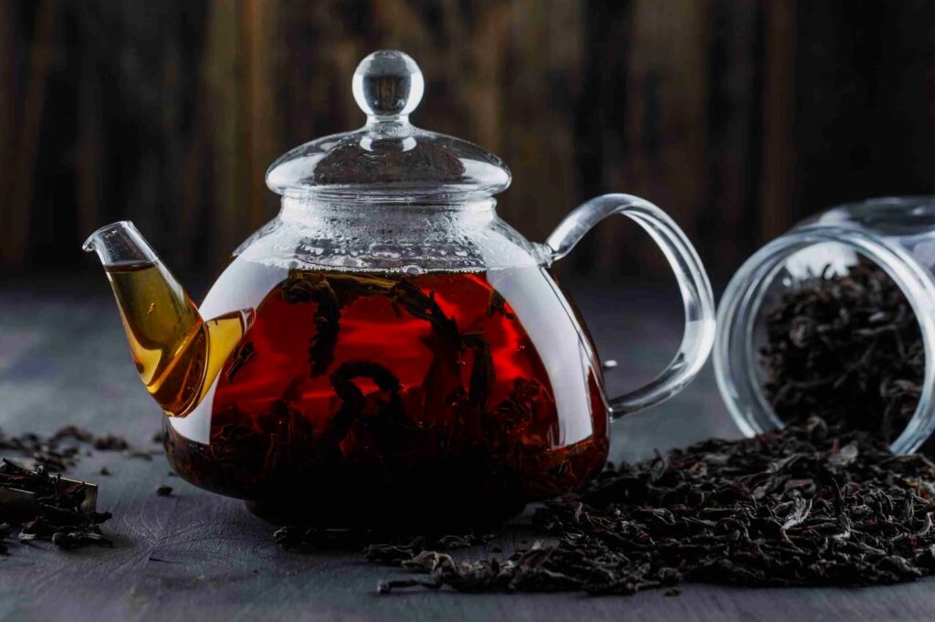 A clear glass teapot filled with dark red tea and leaves, surrounded by loose tea leaves on a wooden surface. A jar lies open in the background. Relaxed mood.