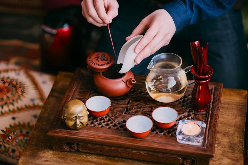 A person prepares tea using a traditional clay teapot on a wooden tray with cups, a small statue, candle, and utensils, creating a calming atmosphere.