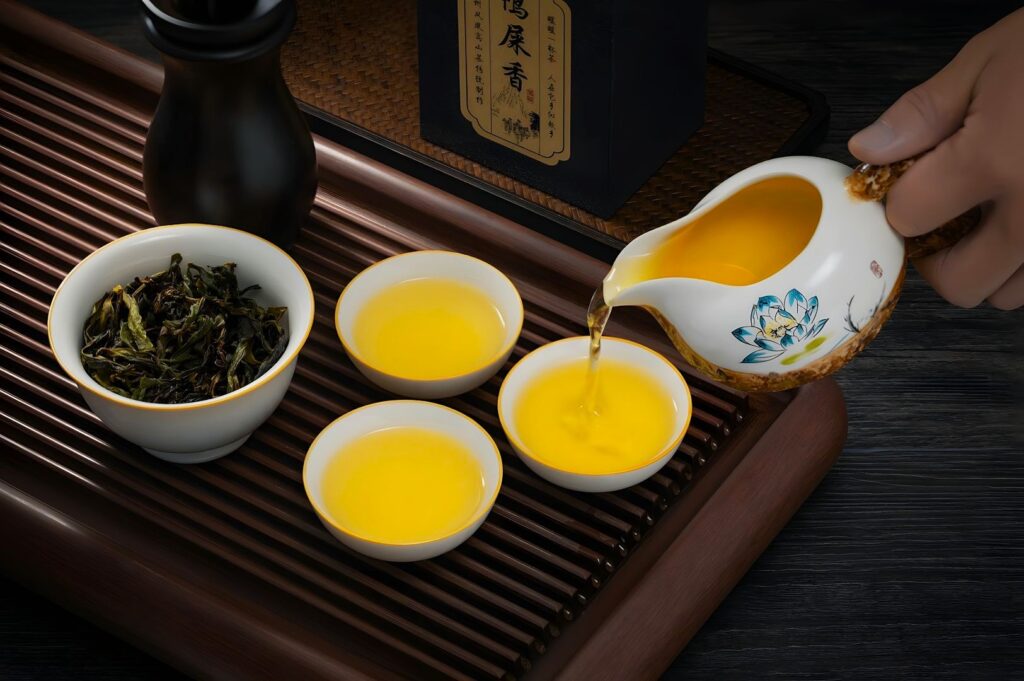 A hand pours golden tea from a decorated teapot into three small white cups on a bamboo tray. A bowl of green tea leaves and a black container are nearby.