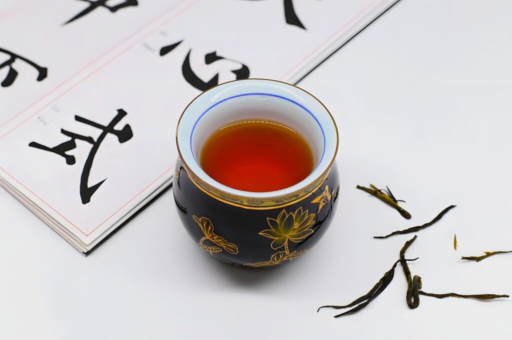 A black cup with gold lotus designs holds amber tea. Next to it are loose tea leaves and an open book with bold black Chinese characters, evoking tranquility.