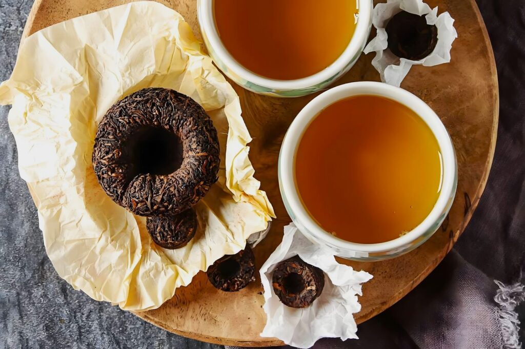 Two cups of amber-colored tea sit on a wooden tray alongside wrapped Pu-erh tea cakes, creating a warm, cozy atmosphere.