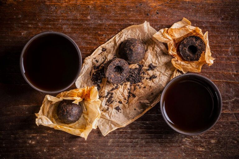 Three dark brown tea pods are wrapped in crumpled paper on a rustic wooden table. Two cups of dark tea are placed on opposite sides, creating a warm, earthy ambiance.