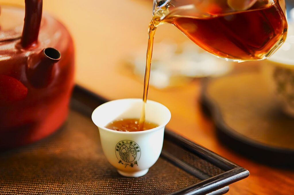 Pouring amber tea into a small white cup with green Chinese characters, beside a red teapot on a wooden tray. Warm and calming atmosphere.