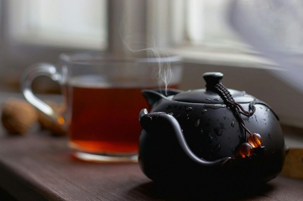 A black teapot with steam and dew sits on a wooden table next to a glass mug filled with dark tea. The scene is cozy, near a softly lit window.