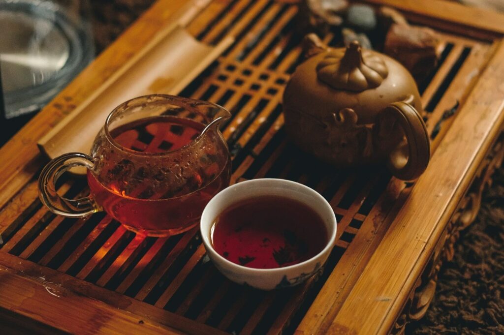 A rustic bamboo tea tray holds a glazed teapot, clear glass pitcher, and a teacup filled with dark amber tea. The scene conveys warmth and tranquility.
