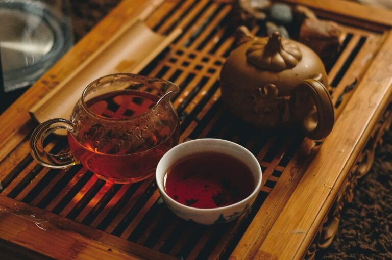 A rustic bamboo tea tray holds a glazed teapot, clear glass pitcher, and a teacup filled with dark amber tea. The scene conveys warmth and tranquility.