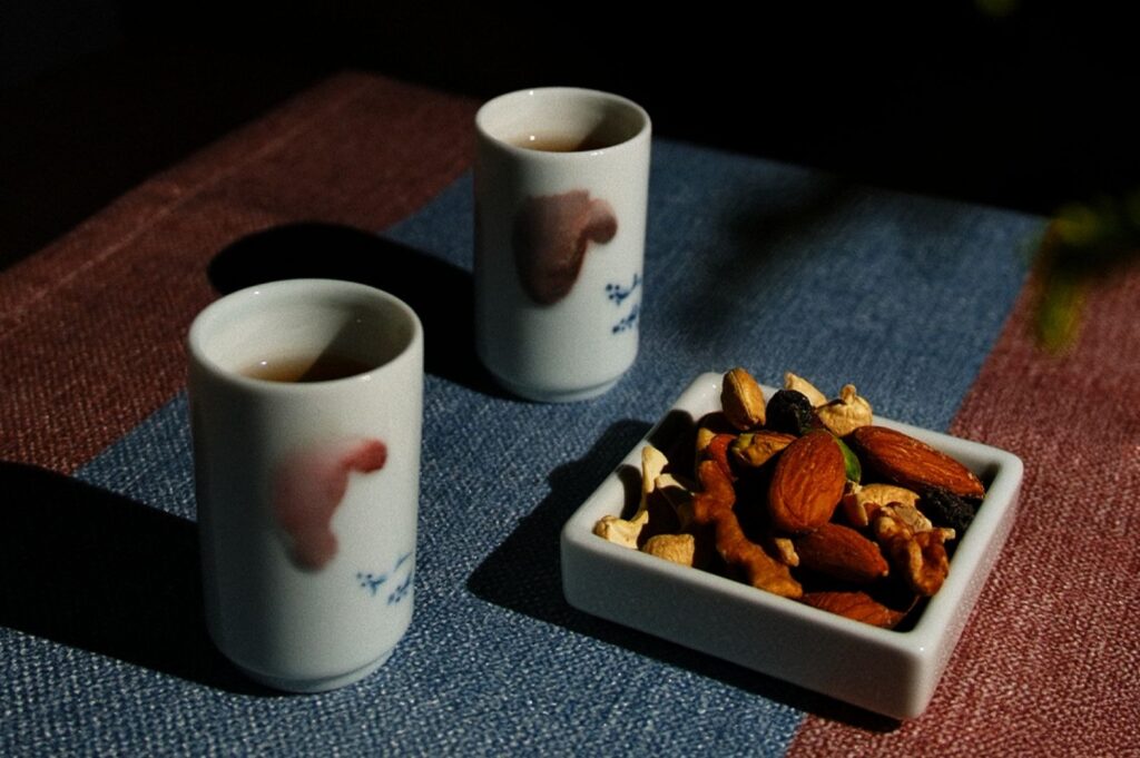 Two white ceramic cups filled with tea and a small square dish of mixed nuts are set on a textured blue and maroon cloth, bathed in warm sunlight.