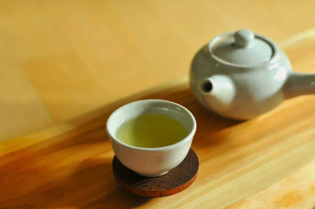 A white ceramic teapot and matching teacup filled with light green tea sit on a wooden tray. The scene conveys a calm and serene atmosphere.