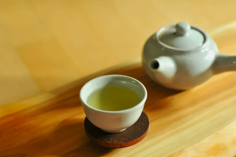 A white ceramic teapot and matching teacup filled with light green tea sit on a wooden tray. The scene conveys a calm and serene atmosphere.
