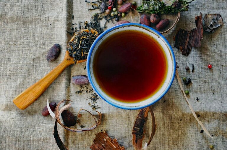 A cup of dark tea on linen fabric, surrounded by loose tea leaves on a wooden spoon, cocoa beans, spices, and bark, creating a rustic and earthy vibe.