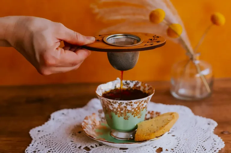 A hand pours tea through a strainer into a floral cup on a doily-covered table. A cookie rests on the saucer, and dried flowers are in the background.