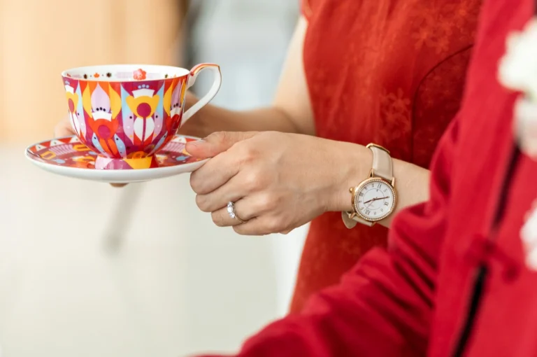 A person in a red outfit holds a colorful, patterned teacup and saucer. They wear a watch and a ring, conveying a sense of elegance and warmth.