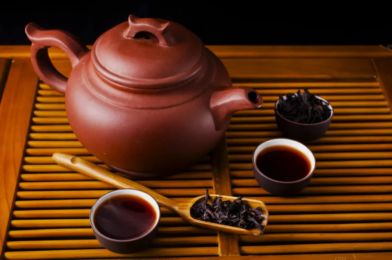 A clay teapot on a bamboo mat, surrounded by two cups of tea and a bamboo spoon filled with loose tea leaves, conveying a warm, calming atmosphere.
