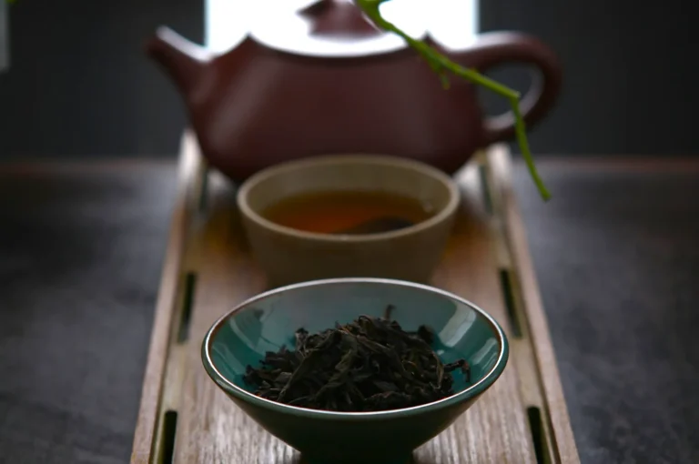 A serene tea scene with a wooden tray holding a teal bowl of loose leaf tea, a cup with tea, and a brown teapot in soft focus, conveying tranquility.