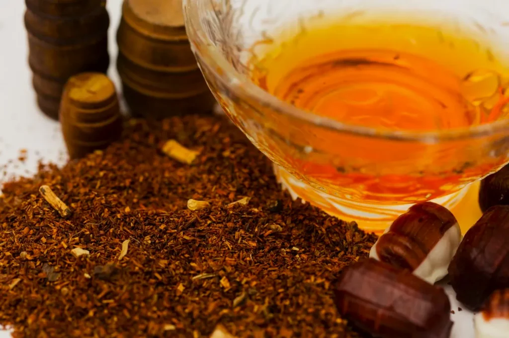 A clear bowl with amber liquid surrounded by loose, dried rooibos tea leaves and stacked wooden discs. The warm tone suggests a cozy, inviting atmosphere.