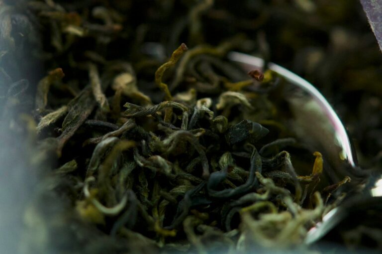 Close-up of loose tea leaves piled in a metal cup, showing curled, wiry strands with dark green-brown tones and subtle silvering.