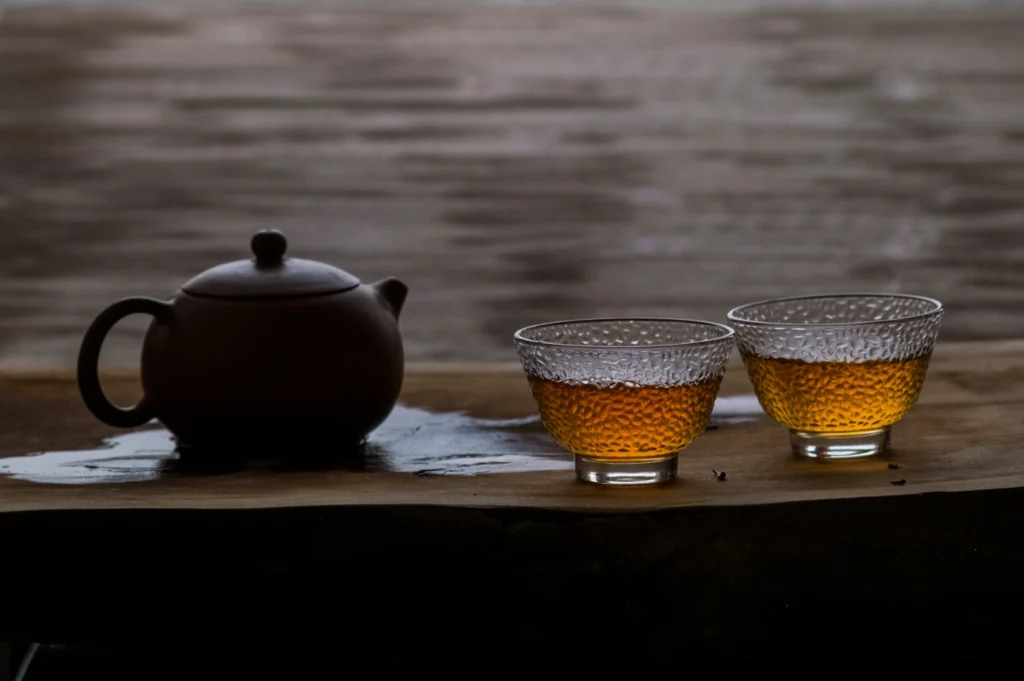 A dark brown teapot and two clear textured glass cups filled with amber tea sit on a wooden surface. A serene and rustic atmosphere is conveyed.